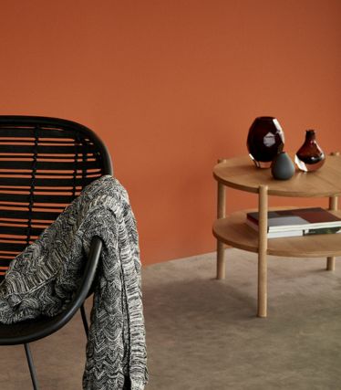 Un interior elegante con la mesa de centro Acron de Hübsch, fabricada en madera clara. La mesa auxiliar redonda se encuentra frente a una cálida pared de color terracota y está decorada con jarrones y libros. Una silla negra con una manta suelta complementa la acogedora escena minimalista