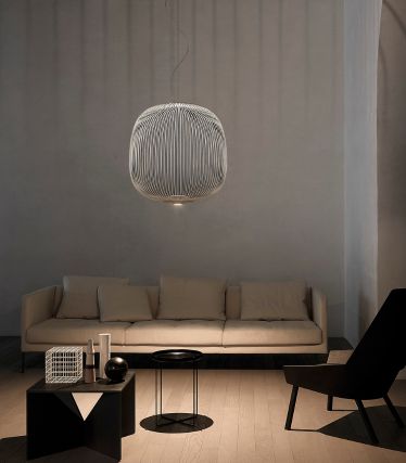Una sala de estar minimalista con la lámpara colgante Spokes de Foscarini como elegante punto focal. Los delicados soportes metálicos de la lámpara proyectan suaves patrones de luz en las paredes y el techo, lo que confiere a la estancia un efecto ligero y escultural. Un sofá de color claro, detalles oscuros y materiales naturales crean un ambiente tranquilo y moderno.