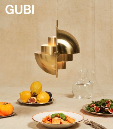 Una elegante lámpara colgante Multi-Lite de GUBI en latón cepillado cuelga sobre una mesa puesta. Debajo hay platos de pasta, higos frescos y limones, así como una jarra de cristal: una escena elegante que combina diseño y disfrute.