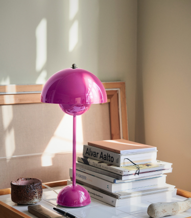 Una lámpara de mesa de color rosa brillante con una pantalla abombada brillante se encuentra sobre un escritorio junto a una pila de libros de diseño, entre los que se incluyen títulos de Alvar Aalto. La suave luz del día ilumina la escena, resaltando la lámpara y proyectando suaves sombras sobre el fondo neutro.