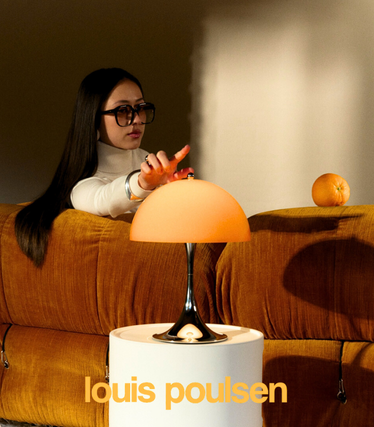 Mujer con gafas de sol toca una lámpara de mesa naranja de Louis Poulsen de la nueva colección Panthella. La lámpara está sobre una mesa auxiliar blanca frente a un sofá naranja, con una luz cálida y suave.