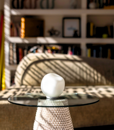 Pequeña lámpara de mesa blanca sobre tapa de cristal, luz solar y estantería al fondo.