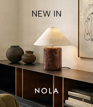 Lámpara de mesa Nola con base de mármol marrón rojizo y pantalla cónica de lino sobre un aparador oscuro. Texto: «Novedad – Nola».