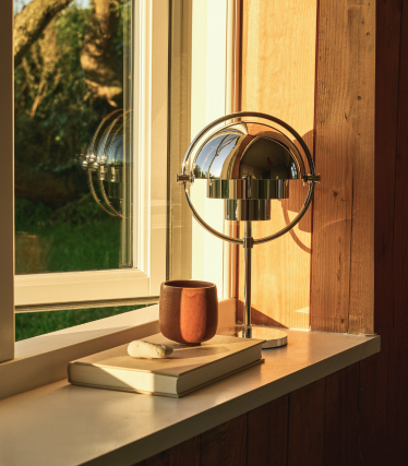 Lámpara de mesa cromada sobre el alféizar de una ventana al atardecer, junto a un libro y una taza de cerámica.