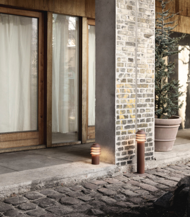 Escena exterior con dos modernas luces cilíndricas colocadas junto a un camino de piedra y una columna de ladrillo y hormigón. Detrás de ellas hay puertas de cristal con marcos de madera y cortinas luminosas, y una gran planta perenne en una maceta. El cálido resplandor de las luces crea un ambiente acogedor y atractivo.