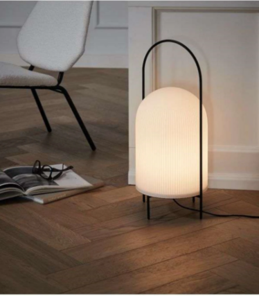 Lámpara blanca acanalada con estructura de asa negra sobre parquet en espiga junto a un sillón.
