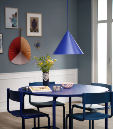 Lámpara colgante azul con forma de cono sobre una mesa de comedor redonda azul con un ramo de flores, en un comedor con pared azul y obras de arte.