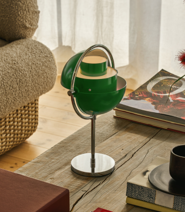 Lámpara de mesa Multi-Lite verde con base cromada sobre una mesa de madera junto a unos libros.